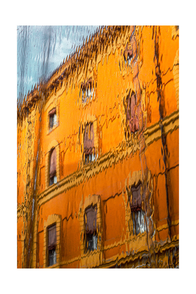 Photograph of an orange-painted building through a plastic rain cover which gives the appearance of peeling paint and visible cracks. The plastic sheet distorts the building with ripples and waves. The building has three rows of windows.The sky in the background is a soft blue, contrasting with the warm, worn tones of the building.
