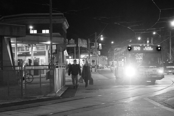 Night. Looking south on Broadview toward Danforth. At left are the lights of the subway station. At right a bus drives toward the camera, its lights flaring. Between them, two people walk toward the camera on the sidewalk with the lights of the busy street corner in the background. 