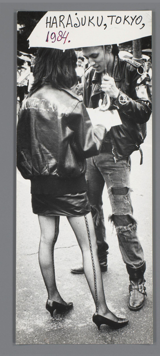 The image captures a moment between two individuals on the bustling streets of Harajuku, Tokyo in 1984. The person standing has shoulder-length hair and is wearing a black leather jacket adorned with white writing that appears to be "ROCK THE" and other unreadable graffiti-like text. They are holding an umbrella over their head, shielding themselves from what could possibly be the rain or strong sunlight. Their attire includes ripped jeans, enhancing the punk aesthetic often associated with Harajuku's street fashion scene.

Beside them is another figure in a black leather skirt that reaches just above the knees and has chains attached to it as part of an unusual accessory. This person also sports high-heeled shoes, creating a stark contrast against their otherwise rugged attire. The background reveals more people walking by on what looks like a market or festival day with various stalls visible.

The overall composition exudes a gritty urban vibe typical of Harajuku's subculture in the late 1980s and early '90s, where fashion often blended elements of punk rock, goth, and streetwear to create bold statements. The presence of Ed van der Elsken’s name suggests this photograph is part of a larger collection or series he documented during his travels through Japan at that time.