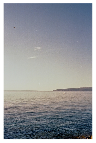 Color photo of the Adriatic Sea with a view of the island of Cres, taken from a swimming spot between Ika and Icici. Although it was taken in the morning, the light is already very bright. The sky takes up almost two-thirds of the image, ranging from blue to almost white on the horizon. A small boat floats in the water. A bird flies into the picture from the left.