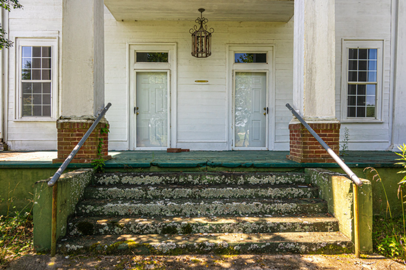 Front entrance of a white wooden house featuring wide concrete steps with green moss growth and lichen, leading to a covered porch supported by tall white columns. The porch has two doors with transom windows above, flanked by symmetrical windows on each side. A decorative wrought iron lantern hangs from the porch ceiling. Metal pipe railings extend from brick pillars on either side of the steps. The structure shows significant weathering and peeling paint throughout. Overgrown vegetation is visible at the base of the stairs and around the foundation."