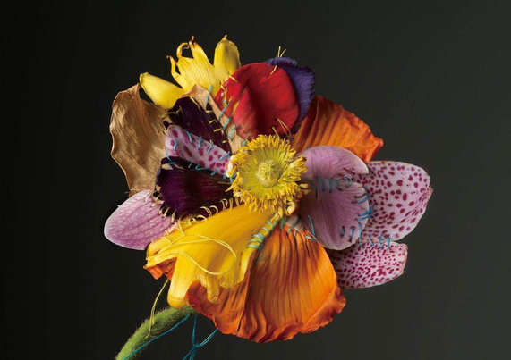 A vibrant, multi-colored flower composed of fabric and petals, intricately stitched together against a dark background.