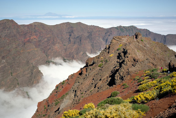 Fotografía de un paisaje montañoso sobre las nubes. A la derecha un camino lleva a un mirador donde se ven dimunutas las personas que estan en el. Vegetacion amarilla y verde contrasta con el rojo de la tierra y el marron de la piedra volcánica. Tras esta masa otra estribacion montañosa aparece sobre las nubes cruzando el cuadro de izquierda a derecha. Sobre esta otra vez una franja de nubes sobre las q se aprecia una franja del oceano, la linea del horizonte y a la izquierda, la isla de Tenerife con el Teide despuntando. Sobre esto el cielo.