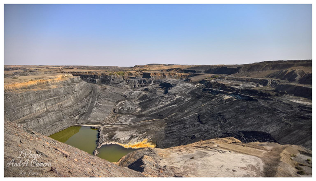 A panoramic, wide angle photograph in colour, signed by Kev Peirce, showing a massive open cut mine.

The image captures the immense scale of the excavation, with distinct, stepped layers (benches) visible along the sides of the pit, highlighting the geology and mining method.

The base of the mine is a mix of dark, excavated material and two pools of stagnant, yellowish green water. The surrounding landscape is arid and flat under a clear, pale blue sky, emphasising the isolation and the environmental impact of the operation.