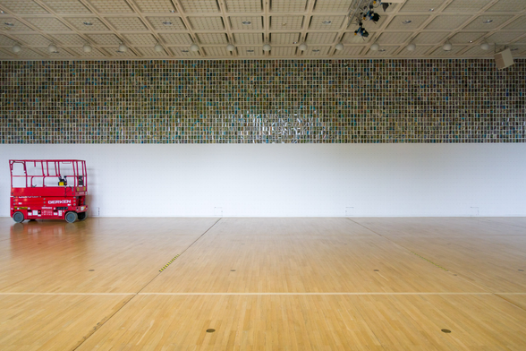 View from outside into the event hall of the Bochum Art Museum. The wooden floor and wall form horizontal lines. A red work platform can be seen on the left. The artwork “No Hiding Place is Completely Safe” by Oriol Vilanova can be seen on the wall.