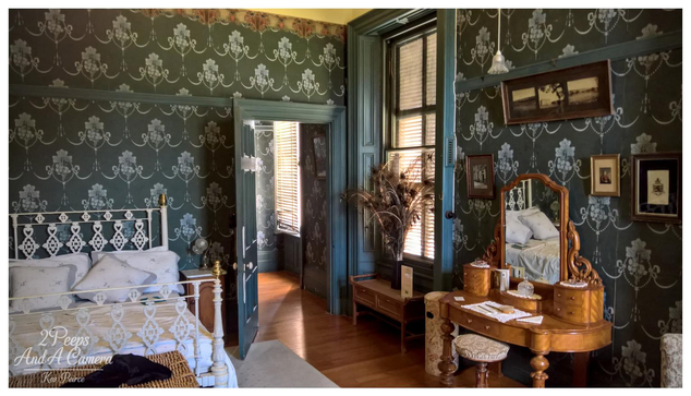 A well preserved Victorian era bedroom at Martindale Hall. The room features dark teal and white patterned wallpaper, polished wooden floorboards, and tall windows with wooden blinds casting long shadows.

On the left is a white wrought iron bed with white linens. To the right, a rich wooden dressing table with an ornate mirror holds various grooming items and reflects the bed.

The door in the centre is open, leading to another richly decorated room bathed in natural light. The photograph is signed Kev Peirce.
