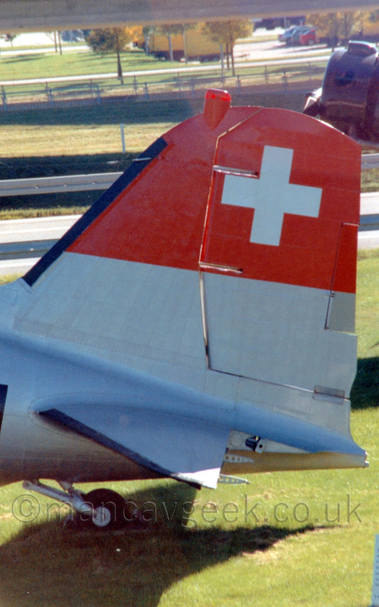 Close-up of the tail of a World War 2-era twin-engined airliner parked on grass facing to the left.
The plane is largely in a silvery-grey bare metal finish, with a thick red stripe at the top of the tail, with a large white cross in the middle, and a large red beacon right at the top.
In the background, the nose/centre engine of a 3-engined airliner peeks into the frame on the left, with grass, trees, and a car park filling the rest of the background.