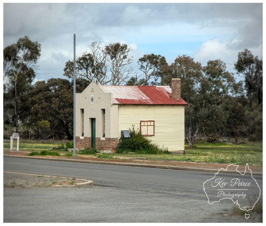 A small, historic looking building in Dumbleyung, Western Australia, with a distinctive design.  The front section is pale grey white with a small brick base and a peaked roof, while the main body is yellow cream corrugated metal with a rusty red corrugated iron roof and a brick chimney.  It sits near a paved road with a light post, surrounded by dry grass and scattered trees under a dramatic, cloudy sky.