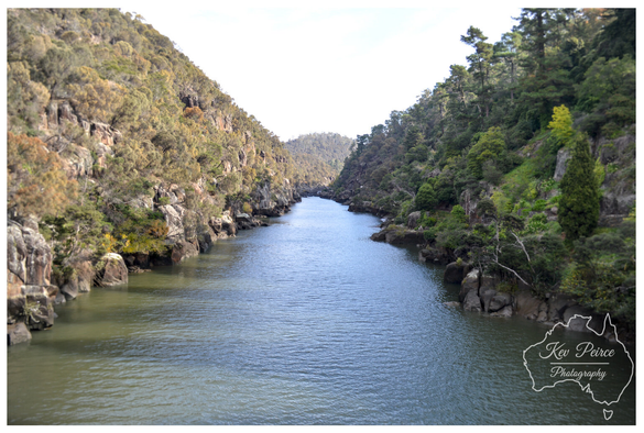 A photograph signed by Kev Peirce showing the Tamar River flowing between two steep, rocky slopes covered in dense green and scrubby brown vegetation.

The river water is a deep blue green, slightly rippled, and fills the narrow gorge. The banks are lined with dark boulders and rocks. The sky visible above the hills is bright white.