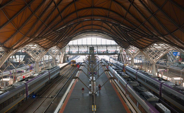 Photo looking down from the first-floor concourse of a major urban railway station. The station has a massive, wave-shaped roof and you can see all the way to the elevated concourse on the other end of the many platforms.