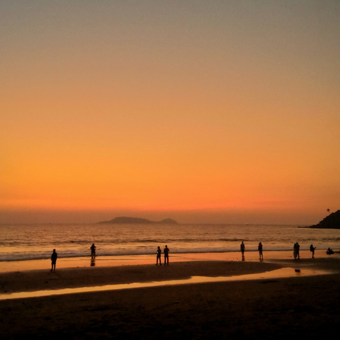 Silhouettes of people stand on a beach at sunset, with a vibrant orange sky and a calm ocean. An island is visible on the horizon.