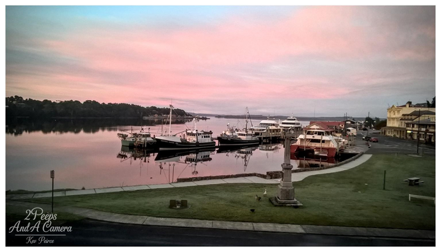 A tranquil, wide angle photograph of the historic Strahan waterfront at sunset. Fishing boats and larger vessels are moored at a jetty reflected in the calm, smooth waters of the harbour.

The sky above is dominated by soft, expansive pink and gold hues. A grassy park and a commemorative stone monument are visible in the foreground. Photo by Kev Peirce.