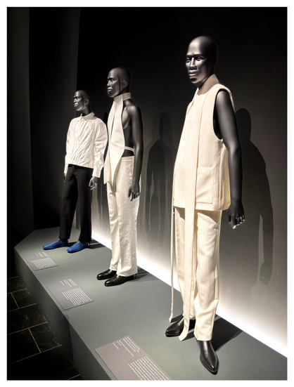 A display of three identical, dark-toned male mannequins wearing different designer outfits in white or cream hues, against a black curtained backdrop. The only spot of color is provided by the blue shoes worn by the mannequin on the far left. Part of the Metropolitan Museum’s exhibition “Superfine: Tailoring Black Style,” about the history and current expressions of Black dandyism.

Designer info from left to right, from the museum placards:

1. Bianca Saunders
Ensemble, autumn/ winter 2024-25
Shirt of white cotton plain weave, trousers of black wool-silk twill

2. Maximilian Davis 
Ensemble, spring/summer 2021
Top of white cotton poplin, trouser of white cotton drill

3. Ib Kamara 
Ensemble, spring/summer 2023
Sleeveless jacket and trousers of cream wool twill