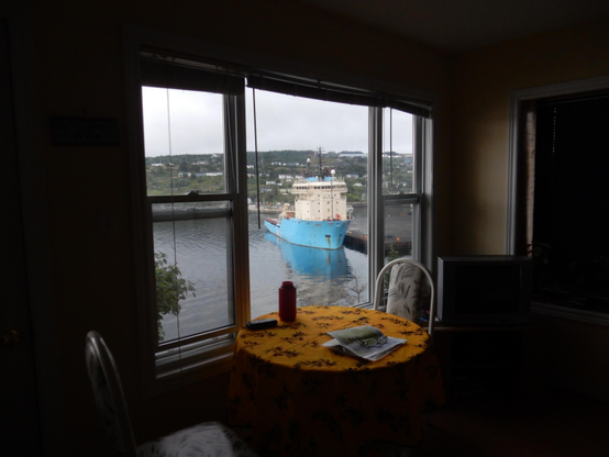 Picture looking across a dark room out a large window. There is a table with yellow table cloth, and two chairs in front of the window, and the view is of a ship with a blue hull in a harbour.