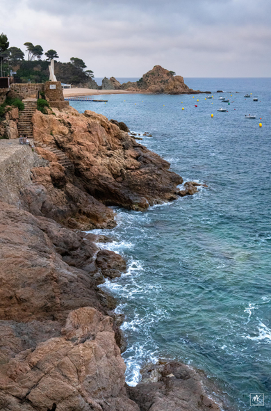 Color photo looking down a rocky coastline with steps coming down the rocks to the water and in the background are small boats moored in the water. 
