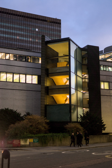 An external staircase with glass walls is illuminated from within, photographed at dusk. A small group of men in winter coats is walking past.