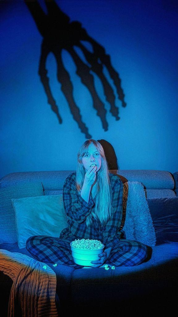 A person in pajamas sits on a couch with a bowl of popcorn, illuminated by blue light, with a creepy skeleton shadow looming behind.