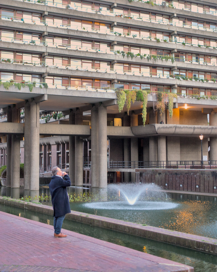 A photo of a man leaning back drinking coffee, looking out over the pool at the Barbican Center, with one of the apartment buildings in the background