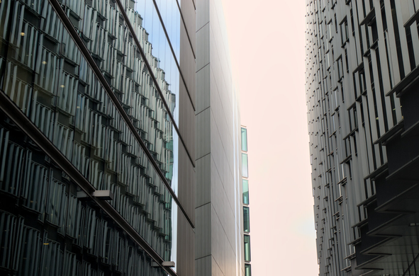A smooth glass facade on the left reflects a facade on the right with repeating metal window frames in a staggered pattern. The hazy sky seen between the buildings is blown out, creating a dreamy effect
