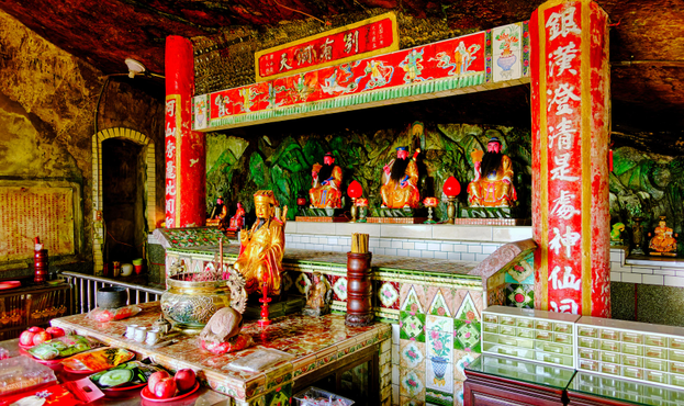Buddhist altar inside a cave with buddha and bodhisattva statues, incense burner, food, fruit, cakes, and a tea set.