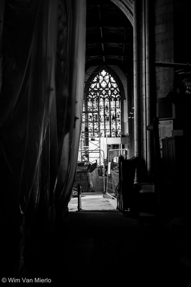 Black and white interior of a church with heavy shadows during construction. Looking in between white sheets draped from ceiling to floor on the left and a stone pillar on the right towards a stained-glass window.