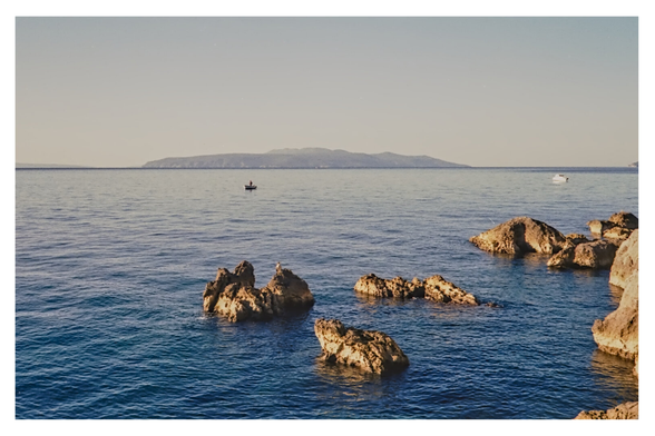 Color photograph of the Adriatic Sea with a view of the island of Cres, taken from a bathing spot between Ika and Ičići. Although taken in the morning, the light is already very bright. The small rocks in the foreground glow bright gold, and the sky ranges from turquois to almost white on the horizon. Two small boats float in the water.