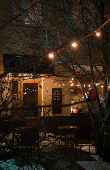The photo shows the entrance and windows of a cozy cafe decorated with garlands of burning light bulbs. Garlands hang on trees. Snow lies on the ground. Late evening.