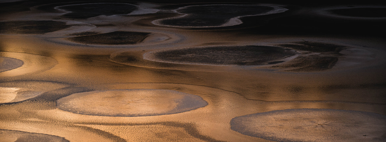 A wide aspect ratio photograph of lake ice. The ice has several large round patterns in it and has a rough surface.

The ice is reflecting the orange sky at sunset, with the topmost part being shadowed