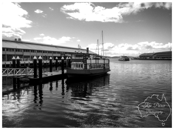 Black and white photograph signed by Kev Peirce, showing a classic moored ferry boat at a dock in Hobart, Tasmania.  The boat is tied to dark wooden posts in the foreground, with its reflection visible in the rippling water. Large, long warehouse-like buildings line the waterfront in the background, under a partly cloudy sky.