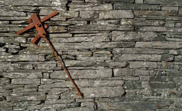 photograph by Remy Dean of a rusted lever mechanism (partial) attached to a grey wall of slate blocks in bright daylight - the arrangement of metal parts resembles Supremacist compositions by the Russian artist Malevich 