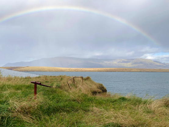 A rusty bendy railing covered with grass and a big rainbow in the sky.

