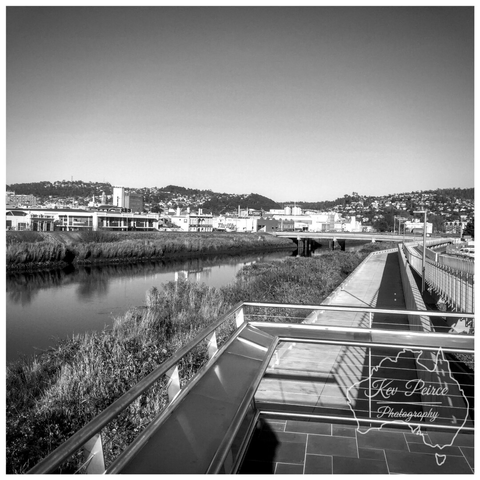 A wide, coloured landscape photograph showing a long, straight gravel path receding directly into the distance between two gently sloping grassy embankments.  The path appears to be on an elevated strip of land. In the background, the houses of Launceston stretch across the low hills under a bright blue sky. A large shadow is cast in the lower right corner.
