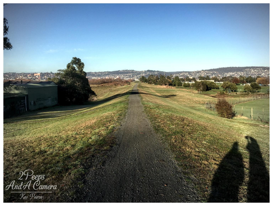 A wide, coloured landscape photograph showing a long, straight gravel path receding directly into the distance between two gently sloping grassy embankments.

The path appears to be on an elevated strip of land. In the background, the houses of Launceston stretch across the low hills under a bright blue sky. A large shadow is cast in the lower right corner.