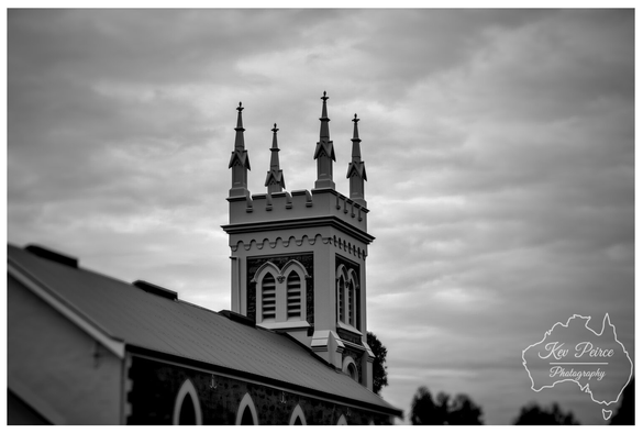Black and white photograph by Kev Peirce showing the crenellated tower and four spire pinnacles of the historic Manaranga Church, with the rough stone church roof leading diagonally into the foreground, all set against a dramatic, cloudy sky.