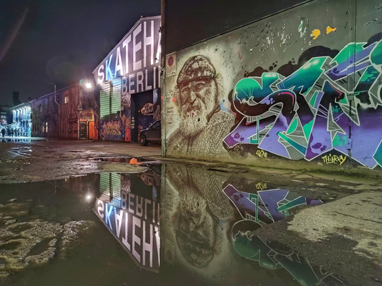 Mehrere große Graffiti an einer Betonwand spiegeln sich nachts in einer großen Pfütze. Im Hintergrund die beleuchtete Skatehalle Berlin.