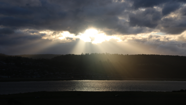 Shafts of light spill through a gap in darkening clouds above a river, casting light rays across a silhouetted hill, through tree tops and over a river