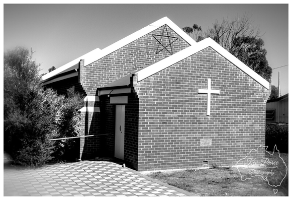 A black and white photograph of a modern brick church building in Dumbleyung, Western Australia. The building features a large white cross emblem mounted on the main facade's brickwork.  Above the cross, set into the white gable end of the slightly taller section of the roof, is a simple outline of the Star of David. A paved checkerboard forecourt is visible in the foreground.