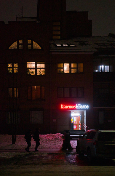 The photo shows a dark, evening street. In the foreground is a house with glowing windows. At the bottom of the house there is a glowing sign of the store, from which a red light falls on the sidewalk covered with snow. In the foreground you can see the silhouettes of cars and people.