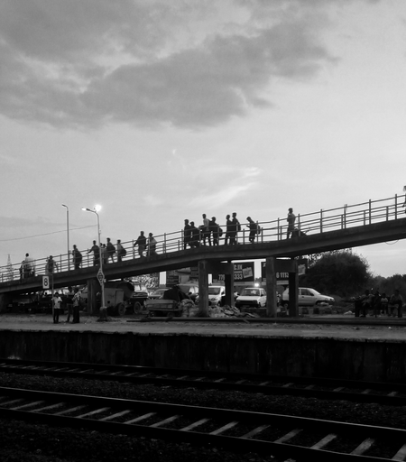 Black and white photo of people walking on an elevated pedestrian bridge at dusk, with parked cars below. Overcast sky adds a moody atmosphere.