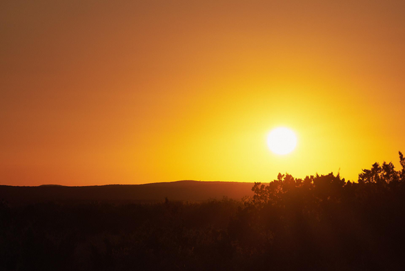 Low sun blazes over a dark, tree-lined horizon, washing the sky in deep gold and orange as heat-hazy light softens the distant rolling hills.