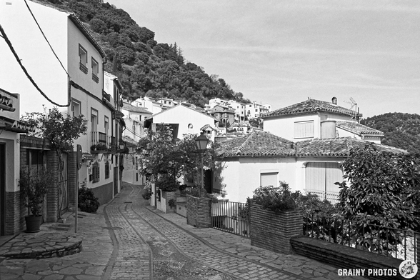 A narrow cobblestone street winds through the picturesque white-washed village of Benelauría, flanked by charming houses and lush greenery, set against a backdrop of rolling hills. The scene is captured in black and white, highlighting the timeless beauty of the architecture.