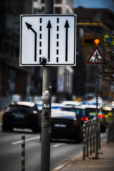 Highly stylized photo of a cityscape that looks like a fever dream, showing a busy city road, taken from the sidewalk. The photo is dizzyingly blurry, except for a high-contrast road lane sign in the foreground. A pedestrian crossing sign is visible behind it.