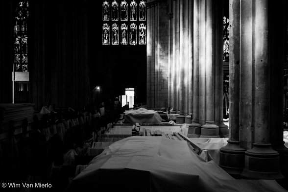 A black and white image of the church interior: the light bounces off the pillars on the right; strong shadows on the left; a stained-glass window in the back. In the centre are various items of furniture covered in white sheets.