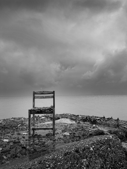 Een versleten/kapotte rieten stoel op blokken beton van een pier in de zee. Op de achtergrond donkere wolken.