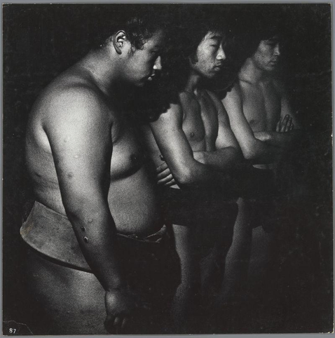 The image is a black and white photograph showing three individuals seated closely together. The person in the foreground appears to be wearing minimal clothing, possibly underwear or traditional attire without sleeves, exposing their shoulders and upper torso. This individual has visible tattoos on both arms. Behind them are two other people with obscured faces; one seems to have short hair while the second's hair is not clearly distinguishable due to lighting and angle.

The image captures a candid moment that suggests an informal setting or possibly a waiting area, given their close proximity and body language suggesting patience or contemplation. The photograph has a grainy texture typical of film photography from its era. There are no discernible background elements apart from the subjects themselves; it is tightly framed to focus on them.

This image's additional information can be found in "Sumo-worshipers, Tokyo" by Ed van der Elsken (1959 - 2010), which was taken during a period when this Dutch photographer explored Japan. The photograph falls under the genre of street photography and documentary work, capturing everyday life with an authentic perspective.

Please note that discussing or making assumptions about individuals in photographs based on appearance can be sensitive; therefore, it's important to focus on objective details without inferring personal characteristics beyond what is visually presented.