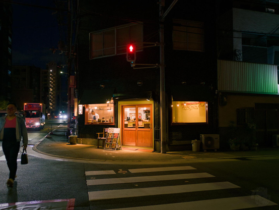 Nighttime urban street intersection. A building with a wooden door and illuminated windows sits at the corner. A pedestrian crosswalk with white stripes spans the road in the foreground. A person walks on the left side, wearing a pink top and dark pants, carrying a bag. A red traffic light is mounted on a pole near the building. In the background, a red truck is visible on the road, with distant buildings and streetlights under a dark sky. The sidewalk features a chair, a signboard, and a trash can.