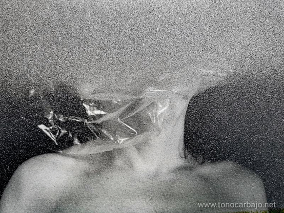La foto en blanco y negro muestra mi cabeza  envuelta en plástico transparente, sobre la fotografía un trazo de spray plateado oculta la parte superior de la foto. Solo queda al descubierto mis hombros, el cuello y laparte superior del pecho.