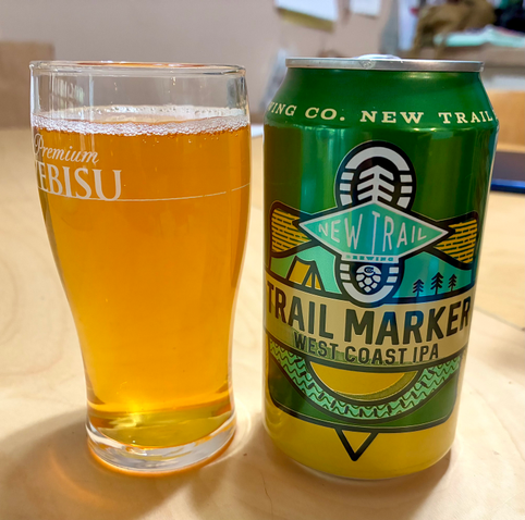 A small Yebisu-branded glass filled with clear, amber-golden beer beside a green Trail Maker West Coast IPA can.