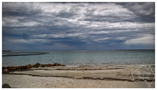 A moody landscape photo of a beach and bay in Wallaroo, South Australia, under a dramatic, dark gray, and heavily clouded sky.  The foreground features pale sand with lines of seaweed, backed by a small cluster of dark rocks. The calm, blue green water stretches out to the horizon, where a break in the clouds hints at a distant downpour.
