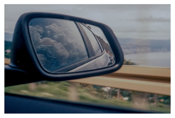 Color photograph of a car side mirror taken from the passenger seat in a moving car. Clouds, the camera, and the two hands holding it are reflected in the mirror. Around the side mirror, the movement of the car causes the roadside guardrail, some sea, and coastline to appear blurred.